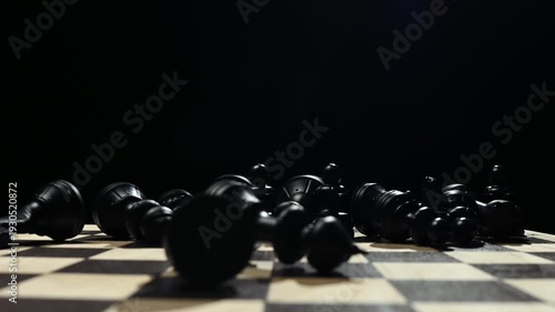 Black chess pieces on a board stand in a row. A man's hand knocks down the pieces on a chessboard on a black background 4к