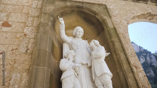A close-up of a white marble sculpture depicting a saintly figure pointing upward while teaching two young children. The statue is set within a traditional stone niche in the Montserrat Abbey complex