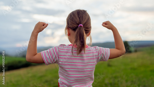 Rear view of a young girl standing outdoors in a green countryside landscape, raising her arms in a gesture of strength and victory. Concept of empowerment, confidence, childhood resilience