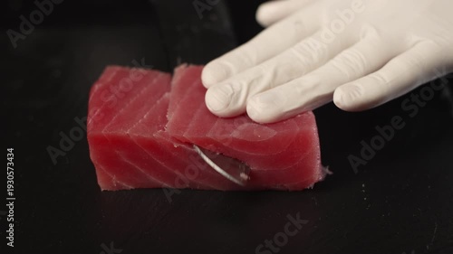 Professional chef with gloves slicing raw tuna fillet with a sharp knife on a black cutting board for preparing delicious sushi, sashimi, or poke bowl in a restaurant kitchen