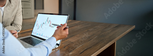 Financial advisor explaining invest stock market data consulting investor. Two business men analysts doing finance trading analysis pointing at chart on laptop screen working in office. Close up.