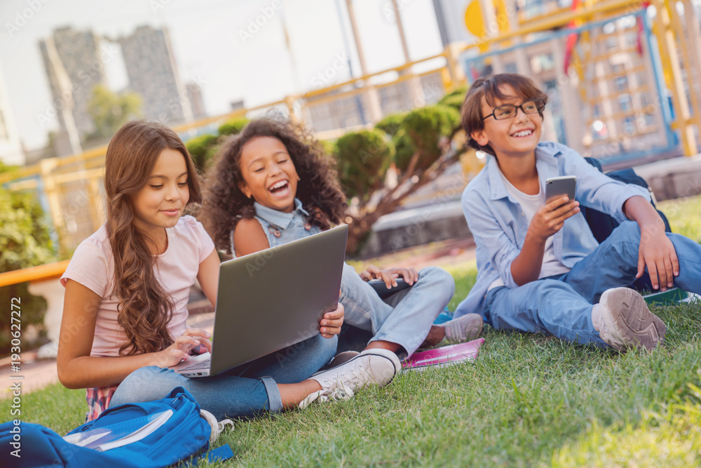 Fototapeta premium Group of smiling multiethnic elementary middle school children pupils classmates sitting on playground grass looking at laptop computer screen doing homework together