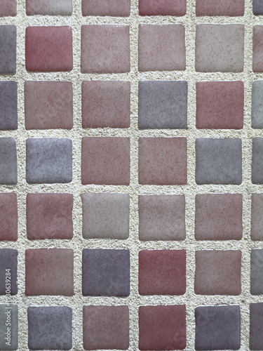 A high-angle macro shot of a decorative mosaic wall surface featuring a grid of square ceramic tiles in muted shades of dusty rose mauve and stone gray set in thick sandy beige grout