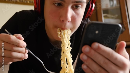 Teenage boy looking at smartphone screen while eating instant noodles with fork, pulling long noodles into mouth. Concept of internet addiction and harm of unhealthy diet.
