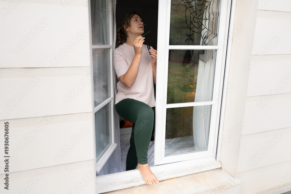 Fototapeta premium Woman painting spiderweb decoration on open window from ledge outside for children