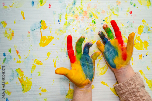 Photo of children’s hands raised palms up, covered in bright paint colors Table surface below is full of paint stains, capturing creativity, artistic play, expressive childhood activity. Copy space