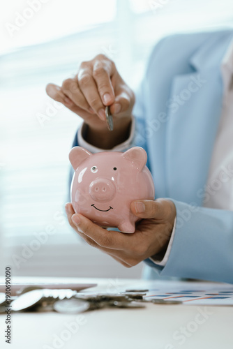 Businesswoman placing a coin into a pink piggy bank on office desk with calculator and charts, representing savings discipline, financial planning, budgeting, and investment growth.