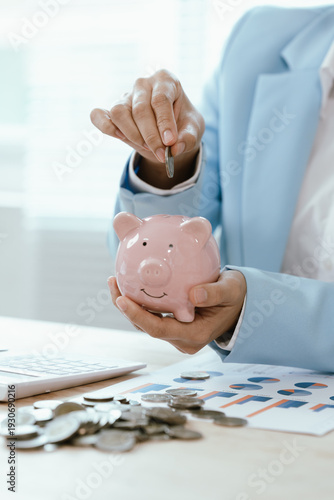 Businesswoman placing a coin into a pink piggy bank on office desk with calculator and charts, representing savings discipline, financial planning, budgeting, and investment growth.