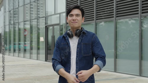 Smiling young Asian man in front of a modern building, casual outfit, headphone 
