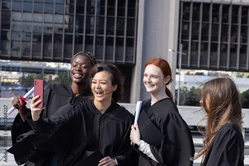 © wavebreak3 - Diverse female graduates in gowns laughing while taking phone selfie at urban plaza with diplomas