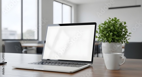 A modern laptop sits on a wooden desk with a potted plant and a white cup