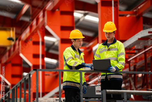 Canvas-taulu Two industrial engineers in safety helmets using laptop on factory platform, discussing production process in modern manufacturing plant with steel structure and safety compliance concept