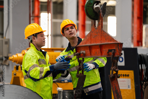Foto Two industrial workers in safety helmets inspecting lifting hook and chain system in factory, checking overhead crane equipment for maintenance and workplace safety compliance
