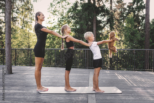 Happy family standing together on yoga mat outdoors in forest park. Mother and children practicing balance exercise with playful dog, healthy lifestyle, active recreation and wellness in nature
