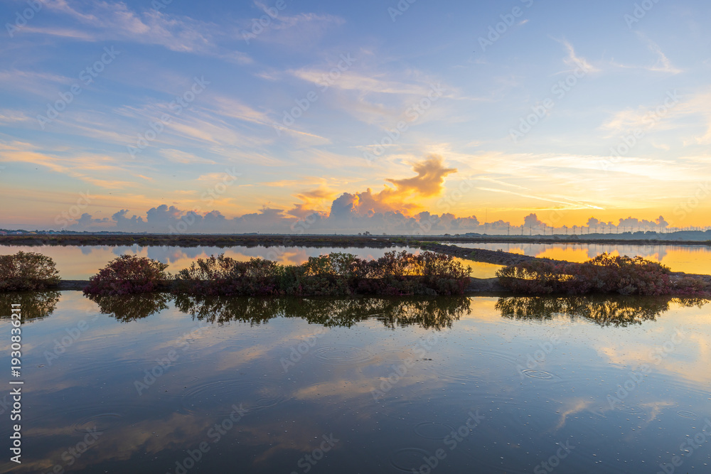 Fototapeta premium Serene Sunrise Reflection Over Salt Fields with Golden Clouds,A peaceful landscape of coastal salt pans or wetlands during a vibrant sunrise. The golden light of the morning sun illuminates dramatic.