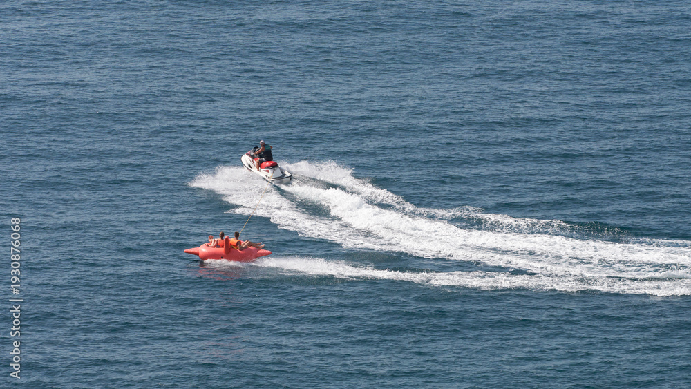 Obraz premium A popular seaside activity is water tubing. A jet ski pulls an inflatable structure with passengers wearing life jackets at high speed.