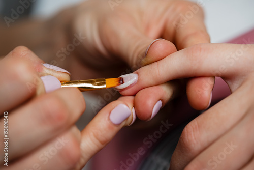 Close up of a professional nail technician using a brush to apply clear gel coating on a female client finger during a manicure service in a wellness spa.
