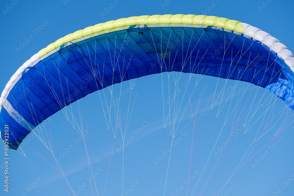 Fototapeta premium Close-up of a paraglider canopy fully inflated against a clear blue sky. Blue, white and yellow wing with suspension lines, paragliding equipment, air sport and freedom concept.