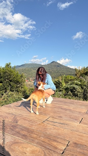 A young woman enjoying a peaceful outdoor moment with her dog on a wooden deck overlooking lush green mountains under a bright blue sky