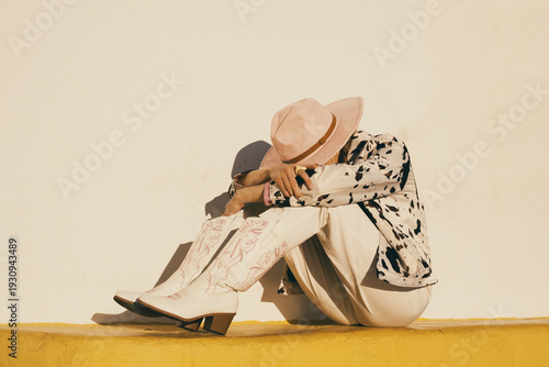 Fotografie Stylish woman in cowgirl outfit sitting against sunlit wall