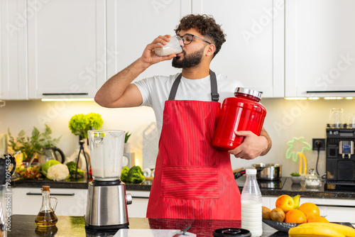 Tableau sur toile Arabian man pours milk whey protein smoothie with banana and apple into a glass after workout in kitchen at home
