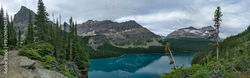 A wide panoramic shot of three mountains and a coniferous forest surrounding a turquoise lake with log cabins on the shore in the Rocky Mountains during summer.