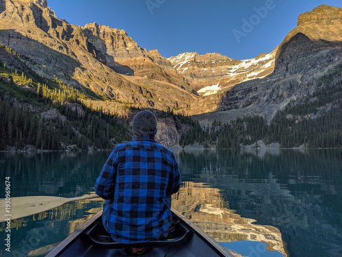 Slika na platnu A man in a blue plaid jacket paddles a canoe towards waterfalls in the Rocky Mountains during sunset