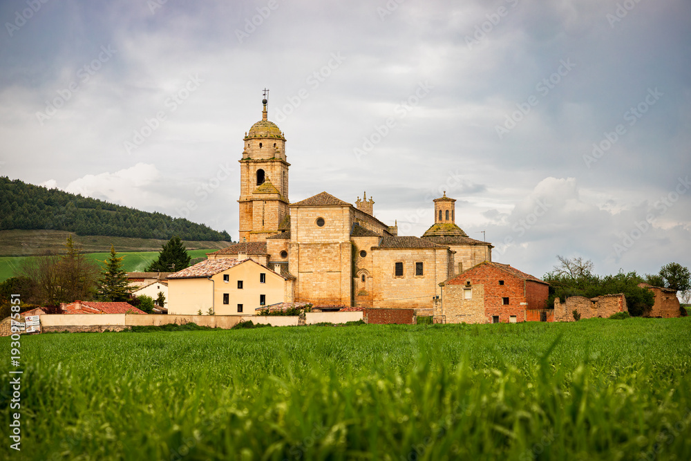 Fototapeta premium Historic stone church and traditional village buildings in rural Spain along the Camino de Santiago, scenic landscape with lush green fields under a cloudy sky in the Spanish countryside.