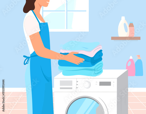 Woman in blue apron holding a stack of clean, folded laundry next to a washing machine in a bright laundry room.