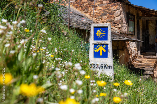 A traditional stone mountain house provides the background for a weathered Camino de Santiago guide marker in Riego de Ambros, identifying the specific route and distance through Spanish countryside.