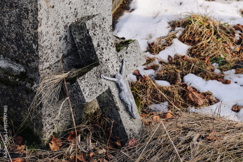 Broken grave cross beside cemetery monument. Snow patches and dry grass in winter. Neglected tombstone detail. Weathered stone symbol. Abandonment and passing time. Forgotten graveyard corner.
