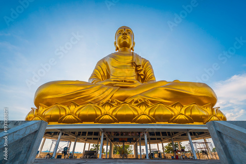 Majestic giant golden Phra Phuttha Lokanat Satsada Buddha statue at Wat Wang Wiwekaram in Sangkhlaburi, Kanchanaburi seated on lotus base beneath radiant blue sky