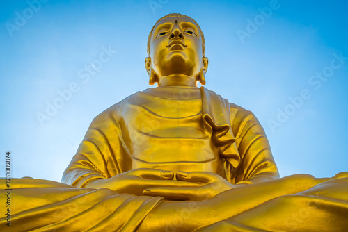 Dramatic low angle view of the giant golden Phra Phuttha Lokanat Satsada Buddha statue at Wat Wang Wiwekaram in Sangkhlaburi, Kanchanaburi beneath clear blue sky