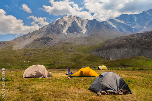 Colorful tents pitched in a high mountain meadow with a massive rocky peak and glaciers above.