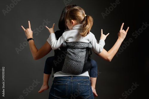 Back view of mother and daughter showing rock and roll hand signs while using a baby carrier.