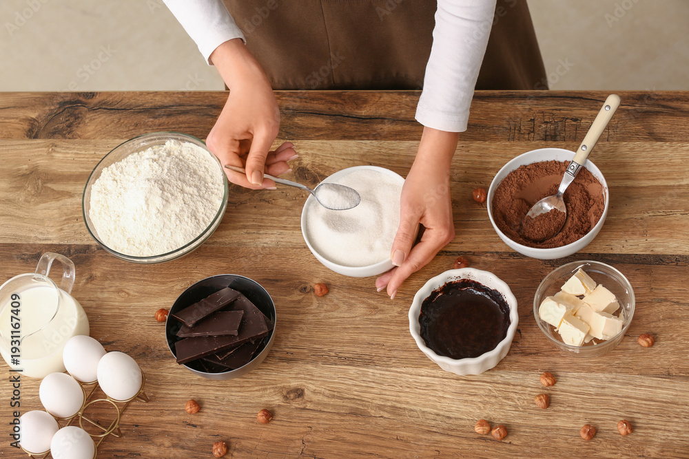 Fototapeta premium Woman adding spoon of sugar into bowl with chocolate for preparing brownie at table in kitchen. Closeup