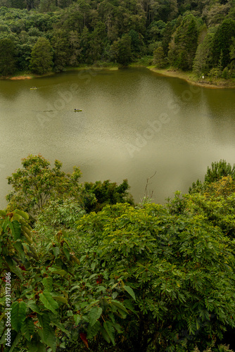 Aerial view of a misty tropical lake surrounded by lush rainforest, scenic mountain landscape with fog rising from trees under a dramatic cloudy sky