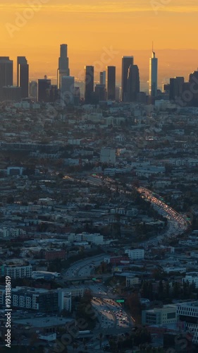 Vertical Screen: Drone aerial view of Downtown Los Angeles skyline at sunrise with heavy morning traffic flowing through freeway toward DTLA skyscrapers in warm golden light, California, USA.