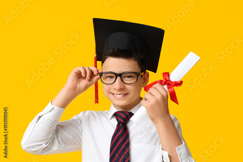 Cute schoolboy in graduation hat with diploma on yellow background
