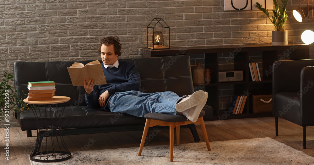 Fototapeta premium Young man reading book on sofa at home late in evening