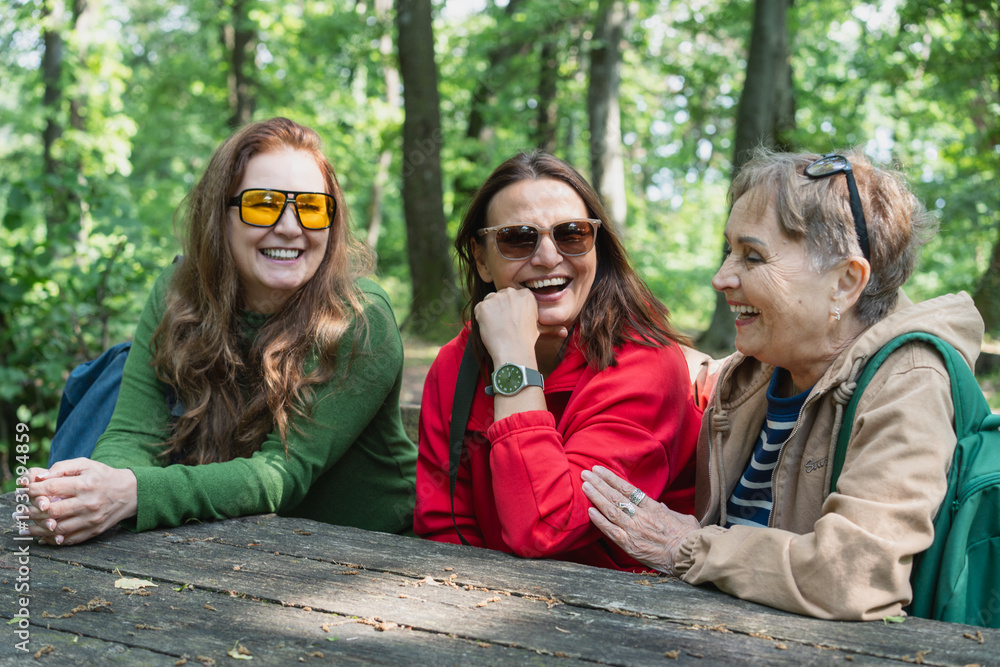 © olezzo - Happy senior mother and two adult daughters sitting at a wooden table in the forest rest area.Happy senior mother and two adult daughters having a picnic at a wooden table in the forest.