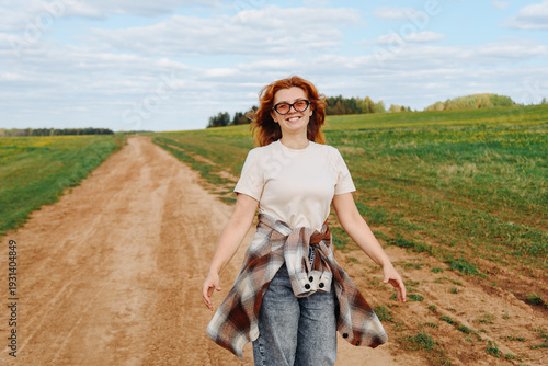 young woman walks on country road, green field, sunny summer day, beige plaid shirt, smiling happily, traveling concept