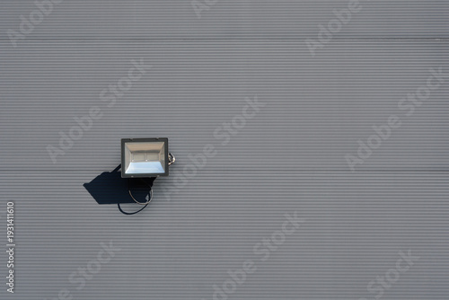 A modern black LED floodlight is mounted on a gray corrugated metal building facade casting a sharp shadow under bright daylight