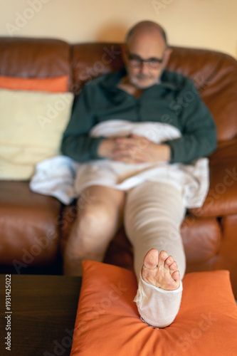 Relaxed elderly man with a bandaged leg resting comfortably on the sofa in his home