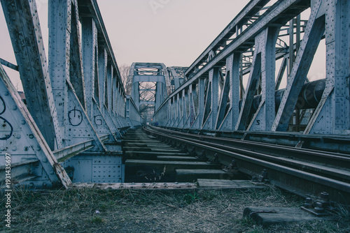 Moody low angle perspective of an old steel truss railway bridge with tracks and wooden sleepers