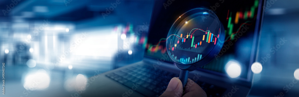 Obraz premium Magnifying glass over candlestick chart and digital financial diagram on laptop, Stock market data analysis and market research, investment strategy and business intelligence data analytics.