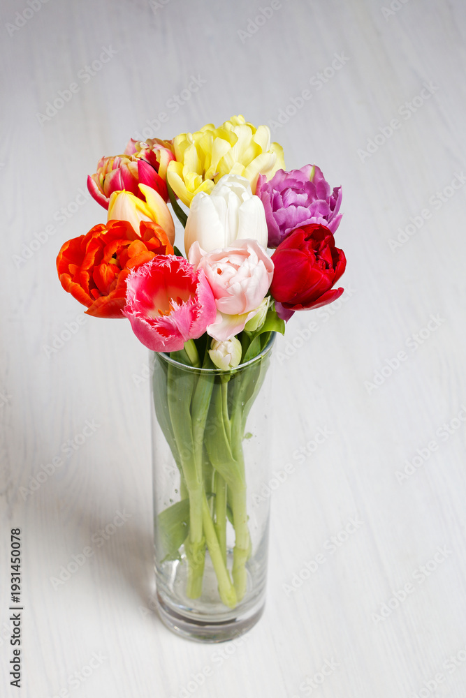 Fototapeta premium Vibrant spring tulips bouquet in cylindrical glass vase on white wooden table. Copy space. Minimalist floral decor for Mother's Day, greeting card or banner design.