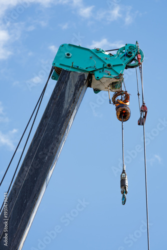 Extended heavy-duty crane boom with teal jib head showcases system of steel cables, pulleys, and suspended hoist hook ready for lifting. Set against clear blue sky backdrop.