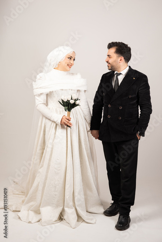 Full-length studio portrait of a happy newly married couple holding hands and smiling at each other. The bride wears a modest white wedding gown with a veil and holds a bouquet of white tulips
