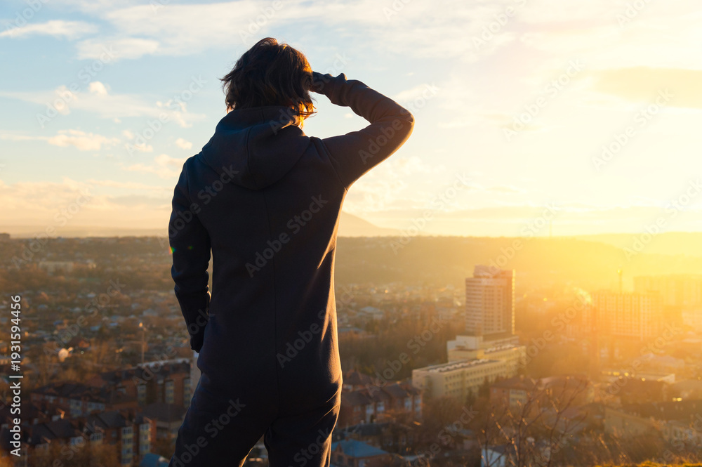 Fototapeta premium Female figure in a fleece jumpsuit stands on hilltop overlooking cityscape at sunset, with buildings and distant mountains visible under a colorful sky